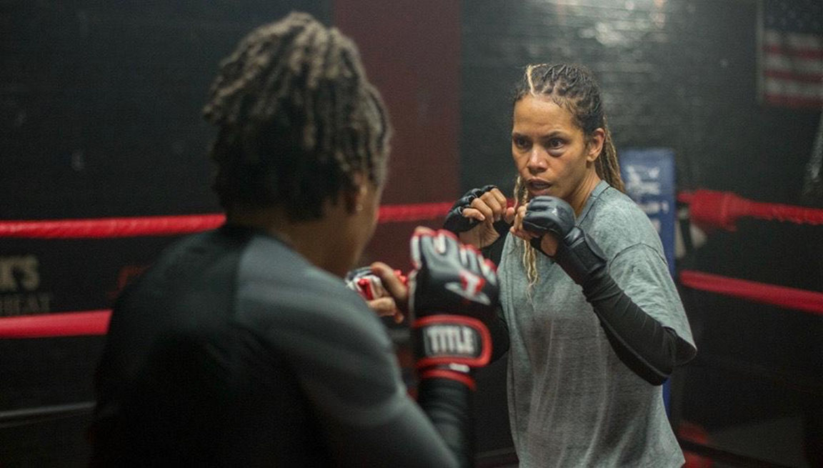 Actress Halle Berry is in a boxing ring as MMA fighter, Jackie Justice, wearing dark gray fighting gloves and a black bruise under her left eye. A publicity shot for the movie BRUISED which she has directed.