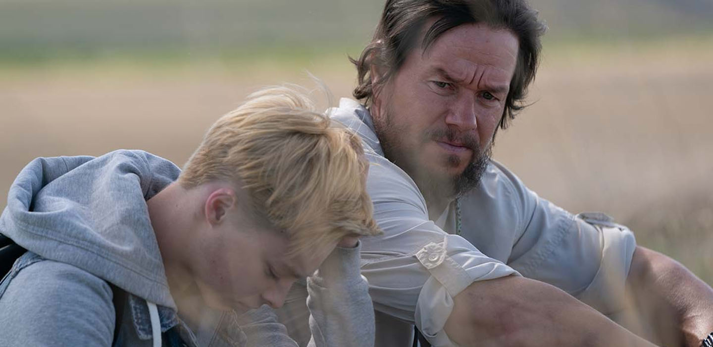 Young actor, Maxwell Jenkins, wearing a light blue hoodie is is sitting down, in the outdoors, next to a bearded Mark Wahlberg (actor) in a publicity photo for GOOD JOE BELL movie