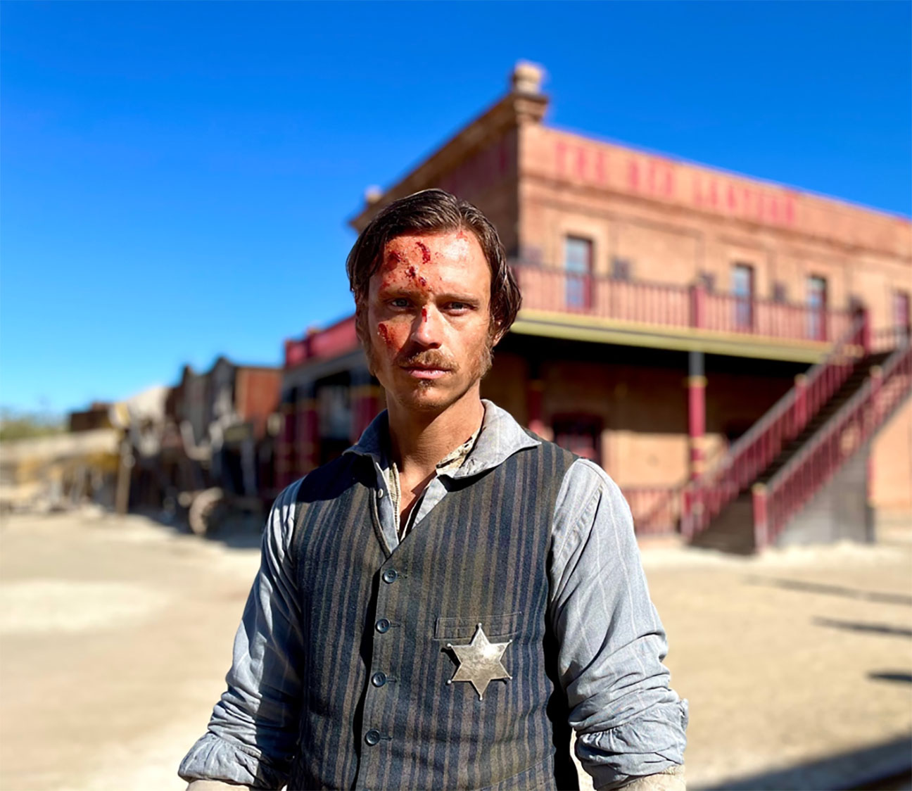 In this photo from the production set, Actor Nicolo Pasetti stanmds in front of an old west building wearing cowboy gear, a tin star on his chest and bruises on his face, on the set of That Dirty Black Bag, upcoming TV series