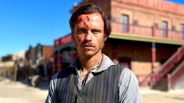 German-Italian actor, Nicolo Pasetti looks bruised in the face as he stands in front of an old building dressed like an old west marshall, wearing a tin star on his chest from the set of That Dirty Black Bag TV series.