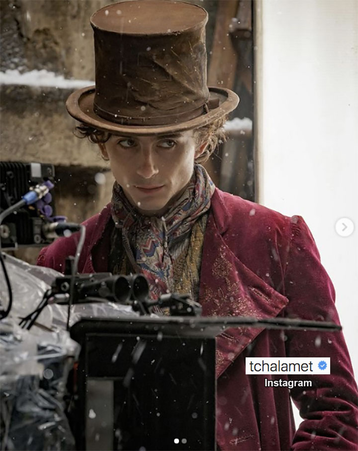 Actor Timothee Chalamet in a brown top hat, scarf, burgundy jacket standing next to a motion camera during the filming of WONKA, a Willy Wonka film.