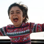 child actor Rayan Sarlak screams in joy with arms open as his upper body sticks out of the sunroof of a sedan in HIT THE ROAD movie.