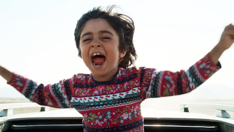 child actor Rayan Sarlak screams in joy with arms open as his upper body sticks out of the sunroof of a sedan in HIT THE ROAD movie.