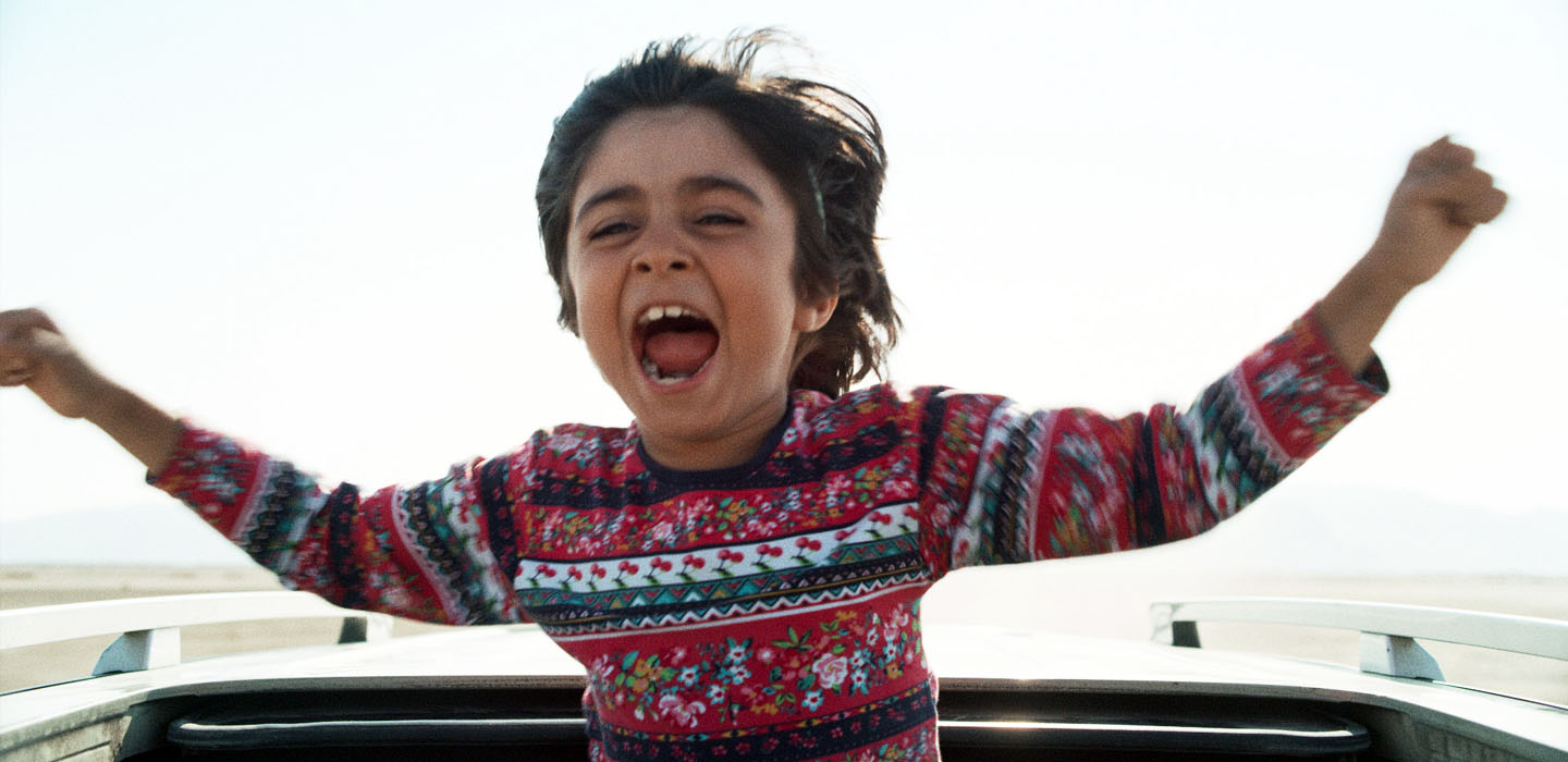 child actor Rayan Sarlak screams in joy with arms open as his upper body sticks out of the sunroof of a sedan in HIT THE ROAD movie.