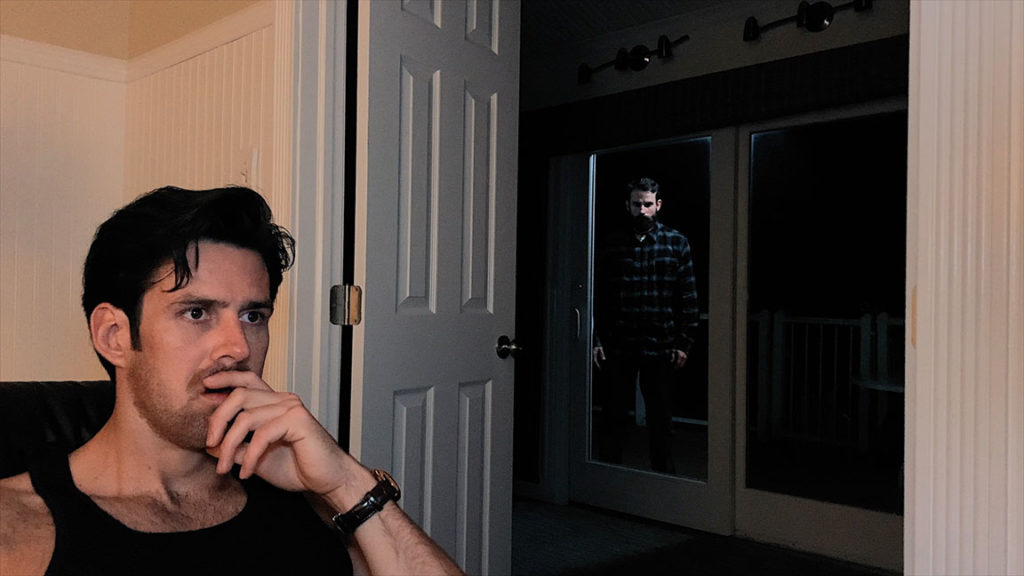 actor, director Bret Lada in a black tank top looking off screen as actor Dustin Fontaine stands outside the glass doors looking at him, in this still phpto from THE ANDY BAKER TAPE film.