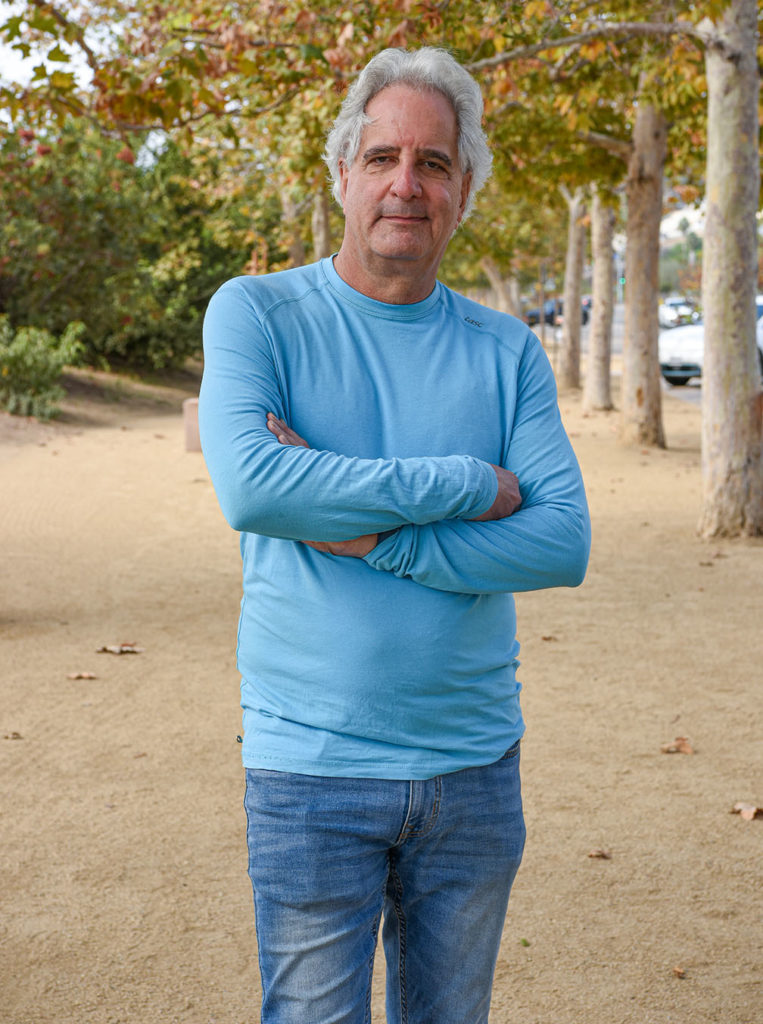 Film producer Michel Shane posing, by a stretch of trees in the background, arms crossed, wearing a longsleeve blue shirt, and blue jeans.